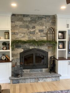 Indoor fireplace constructed with stacked natural stone veneer in varied gray and brown tones, featuring a large black arched fireplace insert, a matching stone hearth, and white built-in shelving units on either side.