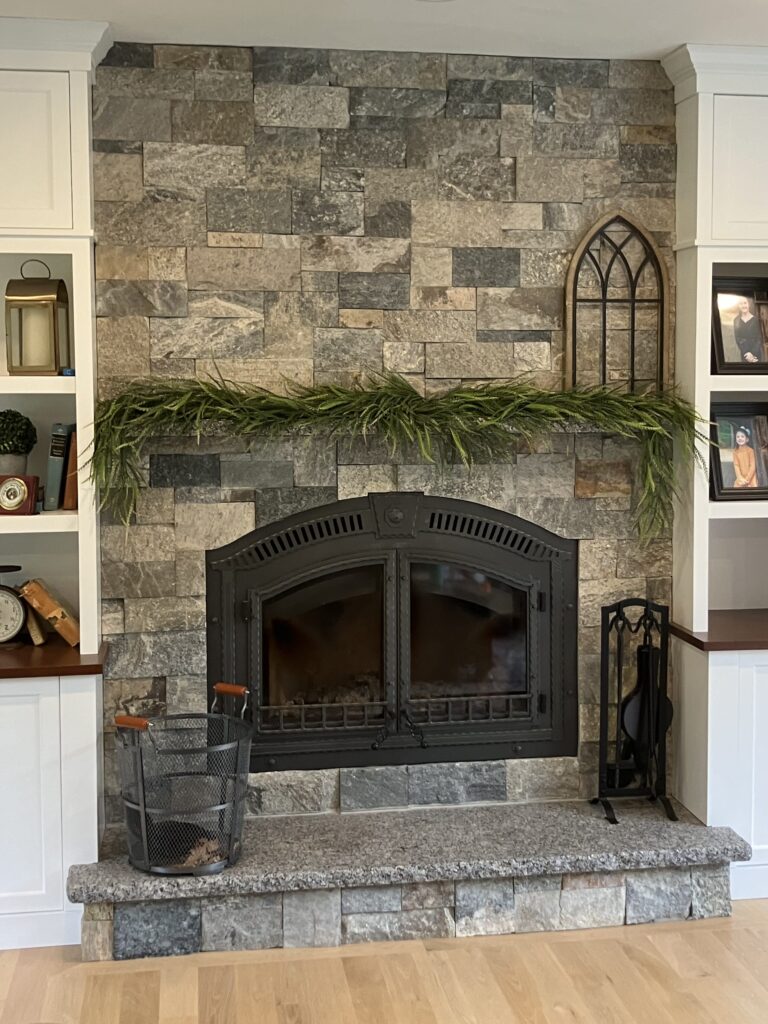 stacked-stone-arched-fireplace Indoor fireplace constructed with stacked natural stone veneer in varied gray and brown tones, featuring a large black arched fireplace insert, a matching stone hearth, and white built-in shelving units on either side.