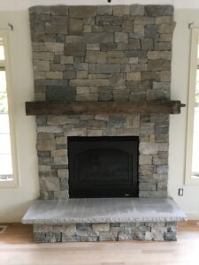 Modern indoor fireplace built with stacked gray and tan stone veneer, featuring a black firebox, a rustic natural wood mantel, and a wide, smooth light-colored stone hearth.