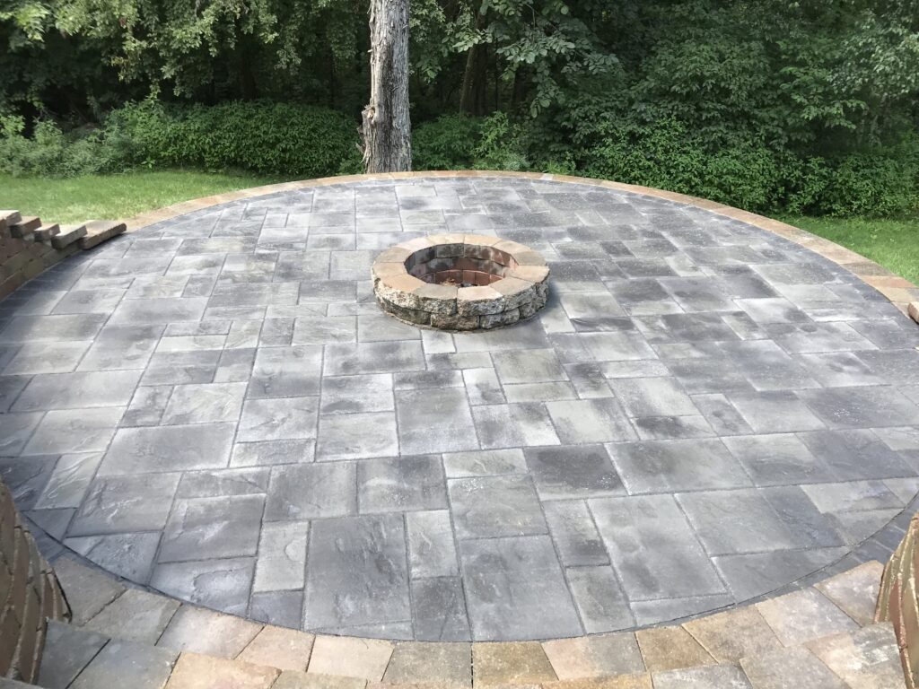 circular-paver-patio-fire-pit Circular outdoor paver patio constructed with varied gray and charcoal pavers, featuring a central stone fire pit and a lighter-toned paver border.