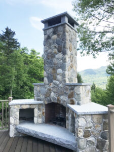 A sturdy fieldstone column topped with a gray granite slab and a traditional lantern-style light fixture, standing in a green grassy yard with trees and a basketball hoop in the background.