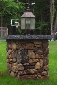 A sturdy fieldstone column topped with a gray granite slab and a traditional lantern-style light fixture, standing in a green grassy yard with trees and a basketball hoop in the background.