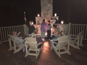 A group of people gather around an outdoor stone fireplace on a deck at night, with some roasting marshmallows over the fire, and Adirondack chairs arranged around the warm glow.