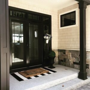 Modern home entrance with black double doors and sidelights, a white-trimmed window, light shingle siding, and a stone veneer foundation.