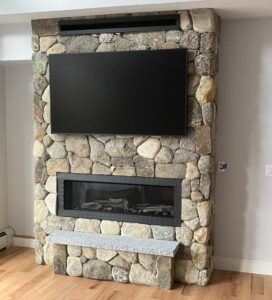 Large, modern indoor fireplace constructed with irregular natural fieldstones, featuring a flat-screen TV mounted above a linear firebox, and an integrated light-colored stone bench hearth.