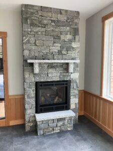 Modern indoor fireplace constructed with stacked gray stone veneer, featuring a black firebox, a light granite mantel, and a matching raised stone hearth, extending high towards the ceiling.