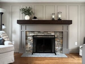 Modern indoor fireplace featuring a stacked gray stone veneer surround, light gray painted wood paneling, and a dark stained wooden mantel, with a black firebox and screen.