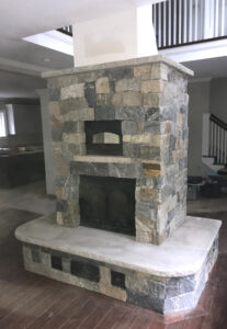 A newly constructed, grand fireplace features a blend of natural stones in various shades of gray and brown, a built-in pizza oven above a large firebox with black doors, and a smooth, light-colored hearth. The fireplace anchors a room with hardwood floors and glimpses of a staircase and kitchen in the background.