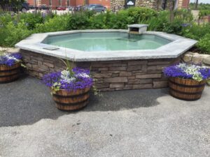 Octagonal outdoor water feature or hot tub built with a stacked stone veneer base and light stone coping, featuring a small waterfall spout and surrounded by vibrant purple and green landscaping with wooden barrel planters.