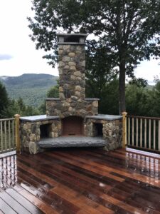 Custom outdoor stone fireplace with built-in wood storage on a polished wooden deck overlooking mountains.