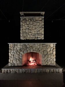 An outdoor stone fireplace with a roaring fire in the firebox, featuring a large river rock base and chimney, set against a dark night sky.