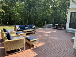 An expansive outdoor patio made of red brick pavers features a wicker lounge set with navy cushions and a dining area with a long table and chairs, all surrounded by lush green trees and a stone wall.