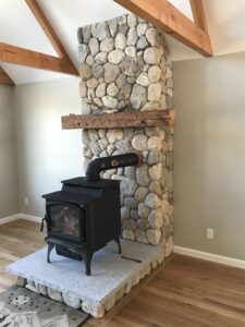 A black wood stove sits on a raised river rock hearth with a light gray carpeted surface, in front of a floor-to-ceiling river rock fireplace with a rustic wooden mantel and exposed ceiling beams.