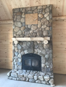 Rustic indoor fireplace constructed with varied river stones, featuring a black arched firebox, a natural log mantel, and a matching stone hearth, set within a wood-paneled room.
