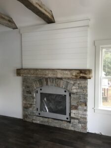 Indoor fireplace featuring a stacked stone surround with an arched opening, a rustic distressed wood mantel, and white shiplap paneling above, with a black metal firebox insert.
