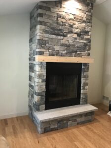 Modern corner indoor fireplace constructed with stacked gray and brown stone veneer, featuring a black firebox, natural wood mantel, and a light granite hearth.