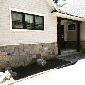 Exterior of a home featuring a light-colored shingle siding and a stone veneer foundation with varying gray and tan stones, next to a black-framed window.