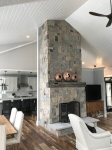Tall, floor-to-ceiling stone fireplace constructed with stacked gray and tan stone veneer, featuring a rustic wood mantel and a large firebox with a screen, serving as a focal point in an open-concept living room and kitchen.